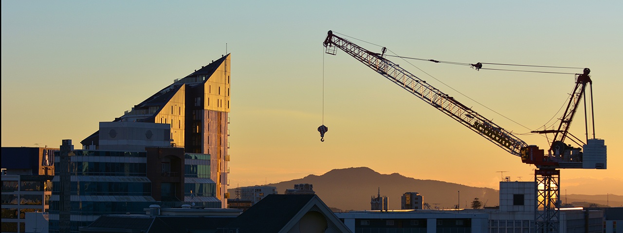 Silhouette of crane and building in Auckland at sunset with Rangitoto Island in background.